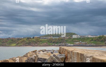 Eine Seeszene mit einem Hügel und weißen Gebäuden auf der Skyline. Ein Pier und Wellenbrecher sind im Vordergrund und dunkle Wolken sind oben. Stockfoto