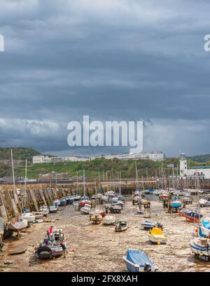 Eine Hafenszene mit Booten in einem Yachthafen und einem Leuchtturm am Ende eines Piers. Ein Hügel mit Gebäuden liegt in der Ferne und schwere Wolken sind darüber. Stockfoto