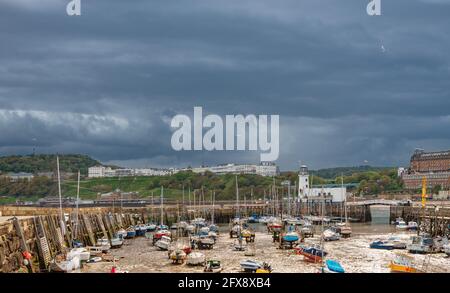 Eine Hafenszene mit Booten in einem Yachthafen und einem Leuchtturm am Ende eines Piers. Ein Hügel mit Gebäuden liegt in der Ferne und schwere Wolken sind darüber. Stockfoto