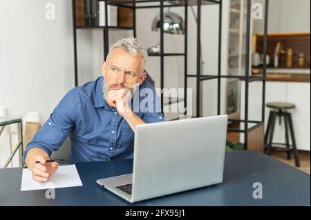 Absorbiert nachdenklichen Senior Geschäftsmann in smart Casual Shirt sitzt am Schreibtisch und mit Laptop, in Gedanken verloren reifen grauen Haar Mann auf der Suche nach neuen Ideen, warten auf Inspiration Stockfoto