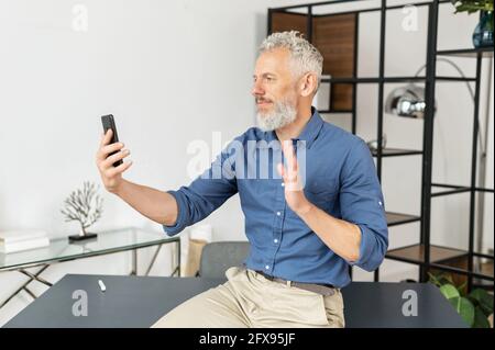 Reifer grauhaariger Geschäftsmann in eleganter Freizeitkleidung nutzt das Smartphone für die Videoverbindung, sitzt in entspannter Pose im Büro, hält Treffen mit Geschäftspartnern ab, spricht mit Kollegen Stockfoto