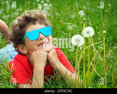 Niedlicher kleiner Junge in blauer Sonnenbrille, der auf dem Gras liegt Mit dem Dandelion Stockfoto