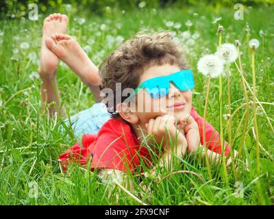 Niedlicher kleiner Junge in blauer Sonnenbrille, der auf dem Gras liegt Mit dem Dandelion Stockfoto