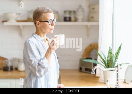 Genießen Sie den neuen Tag. Nachdenklich schöne reife Frau hält Tasse Kaffee oder Tee, reines Wasser, Blick im Fenster und denken oder träumen. Verbringen Freizeit am Sonntagmorgen am Wochenende zu Hause Stockfoto