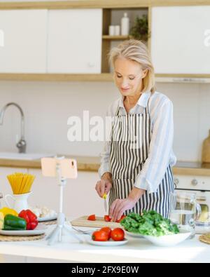 Vertikale Aufnahme von fröhlichen Senior Food Blogger in Vorfeld Aufnahme Video für kulinarische Blog auf dem Smartphone, zeigt den Prozess des Kochens von gesunden Lebensmitteln in der modernen hellen Küche, Schneiden von frischem Gemüse Stockfoto