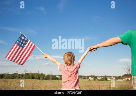 Amerikanische Flagge in der Hand eines Jungen, der mit dem Rücken vor dem Hintergrund des Feldes steht. Halte mit der anderen Hand an der Hand seines Vaters fest. Unabhängigkeitstag Stockfoto