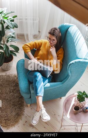 Junge Frau, die Laptop und Kopfhörer benutzt, während sie im Sessel liegt Zu Hause Stockfoto