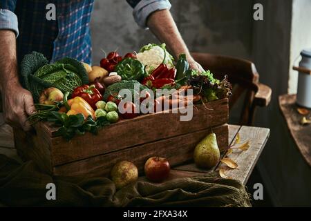 Unkenntlich männliche Putting Holzkiste mit verschiedenen reifen Früchten und Gemüse auf dem Tisch im schmucklosen Zimmer Stockfoto