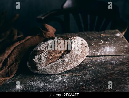 Ganzes Roggenbrot in der Nähe von braunem Tuch aufgelegt Verwitterter Holztisch in dunkler Bäckerei Stockfoto