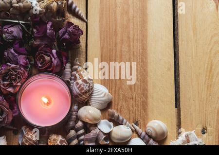 Nahaufnahme einer brennenden rosa Kerze, umgeben von Muscheln, getrockneten rosa Rosen auf einer hölzernen Oberfläche Stockfoto