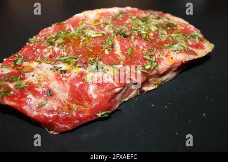 Nahaufnahme von ungekochten Rindfleisch, gewürzt mit Salz, Pfeffer, Basilikum und Zitronenschale, auf einem Schneidebrett aus schwarzem Stein. Stockfoto