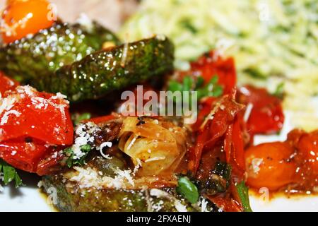 Nahaufnahme von gebratenen Zucchini und Kirschtomaten, mit zerfetztem Parmesankäse, Basilikum und Gewürzen. Orzo und Tuscan Steak im Hintergrund unscharf. Stockfoto