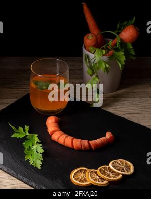 Zusammensetzung von ganzen und auf Rädern Karotten mit geschnittenen getrockneten Orangen Auf Schieferteller und Petersilienblätter und ein Glas Karottenextrakt Stockfoto