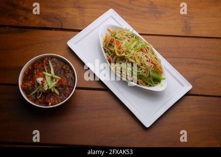 Hühnchen-Hakka-Nudeln mit Chili-Hühnchen-Masala als Beilage Angeordnet in einer ovalen Schale auf weißem Servierteller mit Holzstruktur Stockfoto