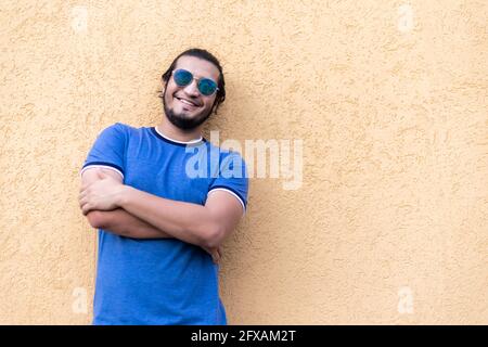 Junger Latino-Mann mit Brille, gekreuzten Armen und langen Haaren, lächelnd und an die Kamera gelehnt. Glückskonzept Stockfoto