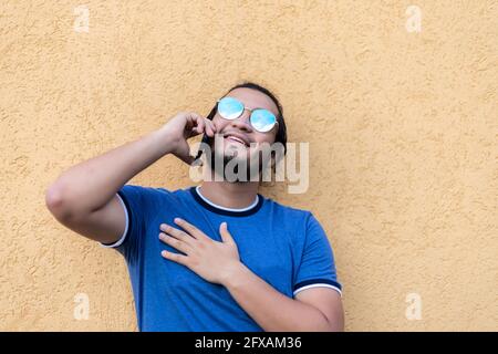 Junger bärtiger lateiner mit Sonnenbrille, die an einer gelben Wand gelehnt ist und am Telefon telefoniert. Frische Luft Stockfoto