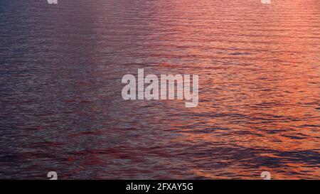 Eine dreifarbige orange, rosa und dunkelblaue Wasseroberfläche mit Wellen und sonnigen Reflexen mitten im Meer. Wasserspiegelung bei Sonnenuntergang oder Sonnenaufgang Stockfoto