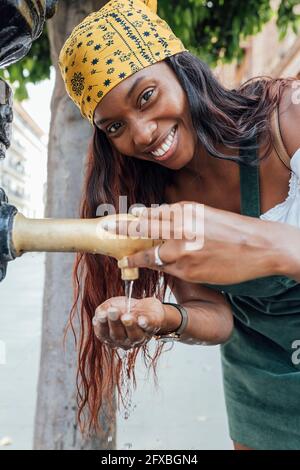 Lächelnde Frau schröpfte Wasser in der Hand durch den Wasserhahn am Trinkbrunnen Stockfoto