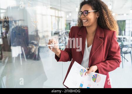 Lächelnde Geschäftsfrau hält den Finanzbericht, während sie im Büro auf Glas schreibt Stockfoto