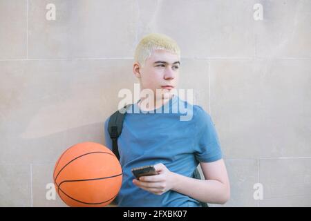 kaukasischer Junge mit Gleichgewichtssball und Handy, gegen das er sich lehnt Die Wand Stockfoto