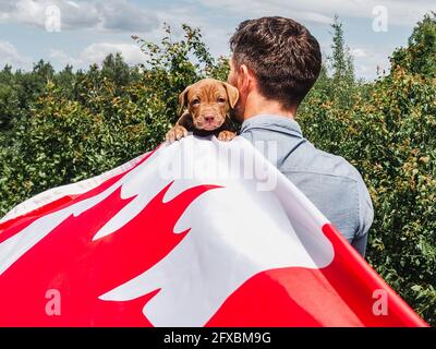 Attraktiver Mann hält einen charmanten Welpen auf einem Hintergrund von blauem Himmel an einem klaren, sonnigen Tag. Im Freien, Nahaufnahme. Nationales Urlaubskonzept Stockfoto