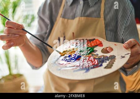 Frau hält die Palette, während sie zu Hause mit dem Pinsel Farben mischt Stockfoto