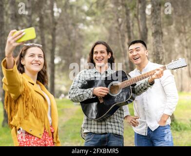 Schöne Frau, die Selfie mit männlichen Freunden beim Picknick im Wald nimmt Stockfoto