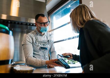 Kundin, die während einer Pandemie über ein Smartphone bezahlt, während sie den Kellner in einer Bar ansieht Stockfoto