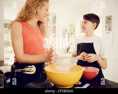 Mutter sietet Puderzucker, während sie den Sohn in der Küche ansieht Stockfoto