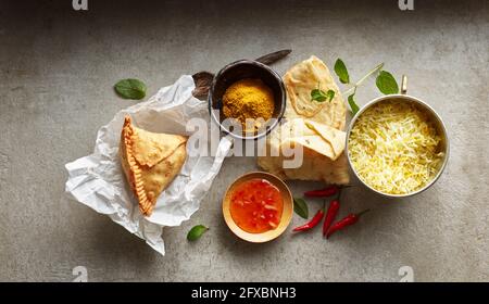 Studioaufnahme von Samosa-Knödel auf Papierserviette, Schüssel mit Basmati-Reis, Chili-Dipping-Sauce, Naan-Brot und roten Chilischoten Stockfoto