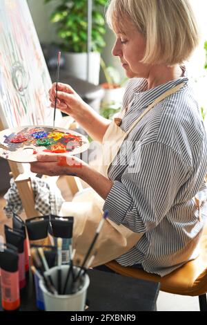 Blonde Frau mit Pinsel, der Farben auf der Palette vor der Staffelei zu Hause mischt Stockfoto