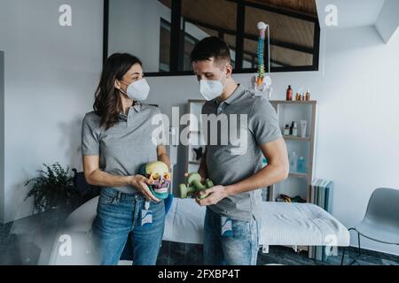 Weibliche Physiotherapeutin mit Schädelobjekt stehend männlicher Kollege, der während einer Pandemie Gewichte vor dem Massageliegen hält Stockfoto