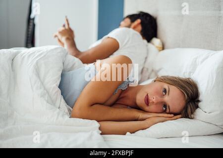 Nachdenkliche junge Frau, die sich hinlegt, während der Mann im Schlafzimmer ein Mobiltelefon benutzt Stockfoto