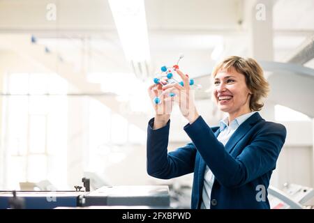 Lächelndes weibliches Fachwissen, das das Molekularstrukturmodell hält, während es in der Fabrik steht Stockfoto