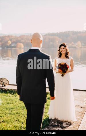 Glückliche Braut, die den Bräutigam ansieht, während sie am See steht Stockfoto