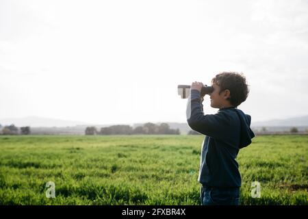 Junge, der während eines sonnigen Tages durch ein Fernglas auf das landwirtschaftliche Feld schaut Stockfoto