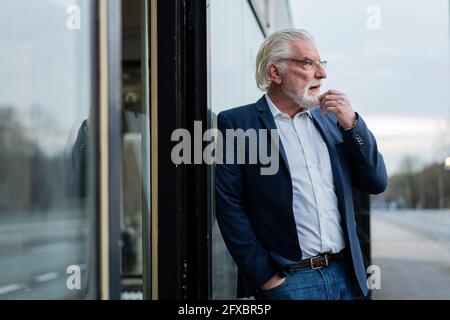 Nachdenklicher Geschäftsmann mit der Hand in der Tasche, die vor dem Büro an der Wand gelehnt ist Stockfoto