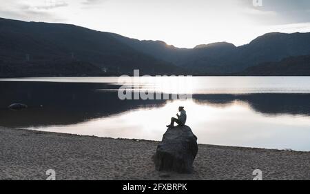 Mann, der während des Sonnenuntergangs auf einem Felsen in der Nähe des Sees sitzt Stockfoto