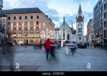 Zufällige Personen, die in der Abenddämmerung auf dem Marienplatz mit dem Alten Rathaus von München, Bayern, Deutschland, auf einem Fußweg spazieren Stockfoto