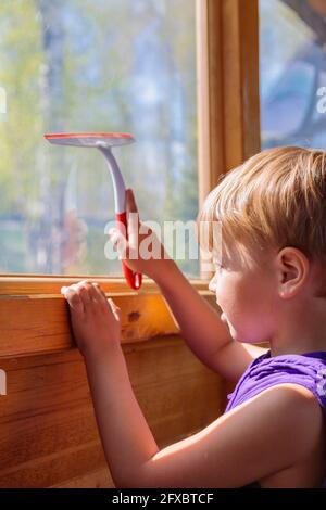 Das Kind wäscht die Fenster mit einer speziellen Bürste. Ein Kind mit blonden Haaren. Stockfoto