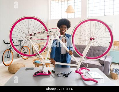 Lächelnde junge Frau, die das Fahrrad auf dem Tisch hält, während sie zu Hause repariert Stockfoto