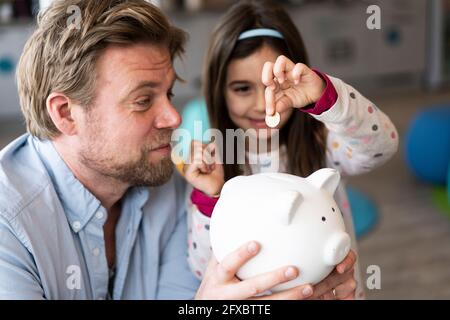 Tochter, die eine Münze in ein Sparschwein legt, das der Vater zu Hause hält Stockfoto