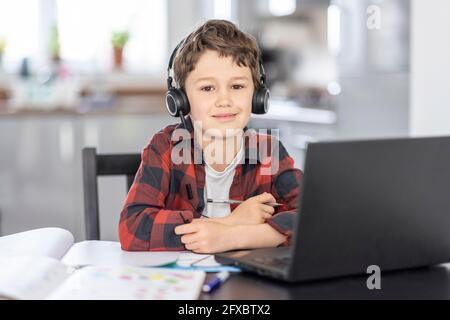 Lächelnder Junge mit Kopfhörern, der zu Hause vor dem Laptop sitzt Stockfoto