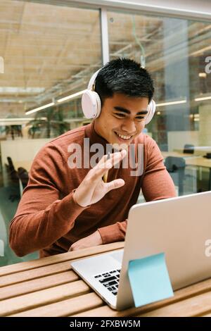 Lächelnder männlicher Unternehmer, der während eines Videoanrufs auf dem Laptop in der Cafeteria die Hand schwenkt Stockfoto