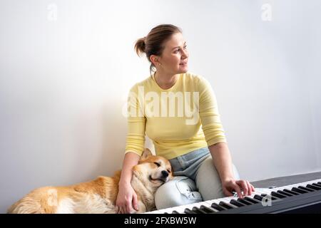 Lächelnde Frau schaut weg, während der Hund auf dem Schoß vor dem Klavier zu Hause schläft Stockfoto