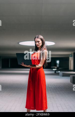 Geschäftsfrau in rotem Kleid, stehend mit Laptop in den Händen in Innenräumen. Stockfoto
