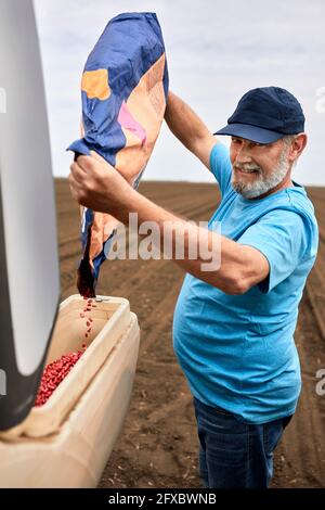 Bärtig reifer männlicher Bauer, der auf dem Bauernhof Maiskörner in einen Behälter gießt Stockfoto