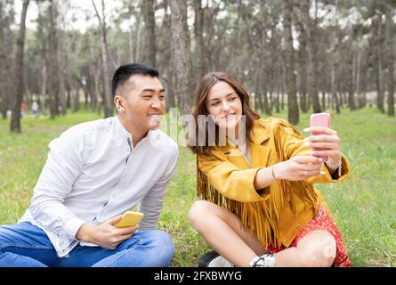 Mann mit einer schönen jungen Frau, die Selfie auf dem Smartphone nimmt, während sie im Wald sitzt Stockfoto