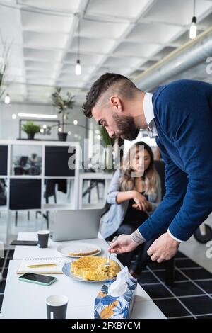 Geschäftsmann, der ein spanisches Omelett am Tisch mit einer Kollegin im Hintergrund im Büro hat Stockfoto