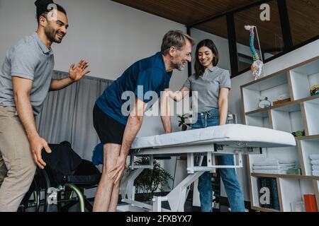 Lächelnde Physiotherapeuten unterstützen den männlichen Patienten beim Aufstehen vom Rollstuhl in der Praxis Stockfoto